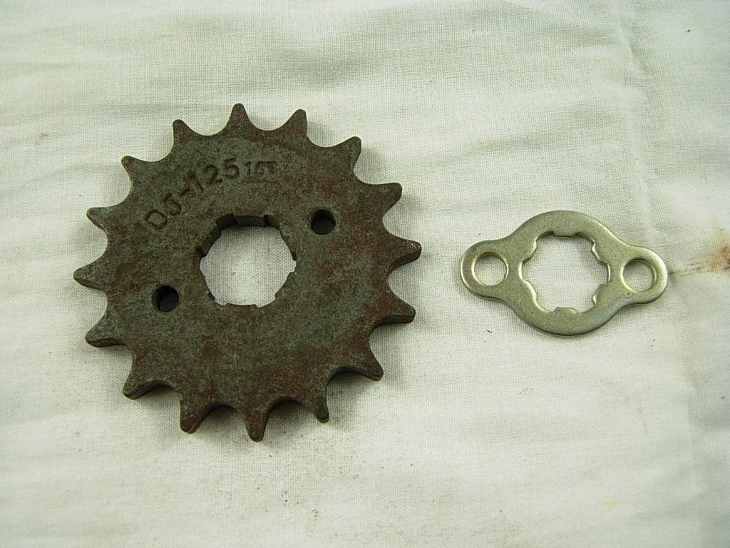 Rusty metal sprocket gear with 12 teeth and a small silver locking plate on a white fabric background