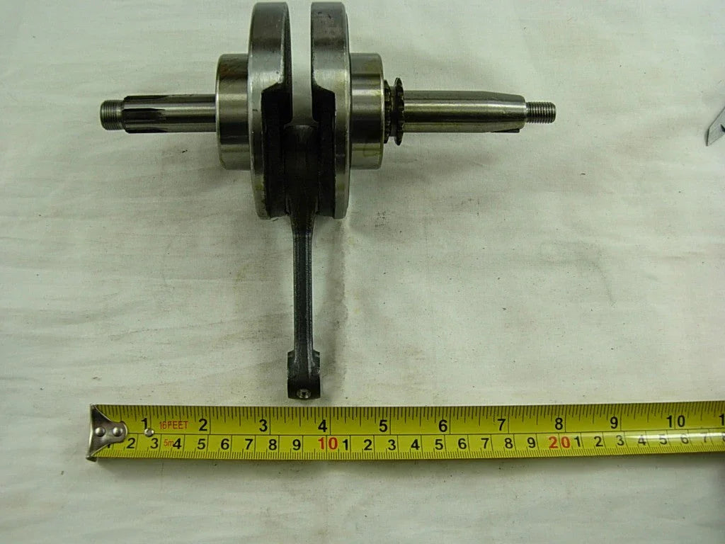 metal crankshaft with connecting rod and sprocket next to yellow measuring tape on white fabric