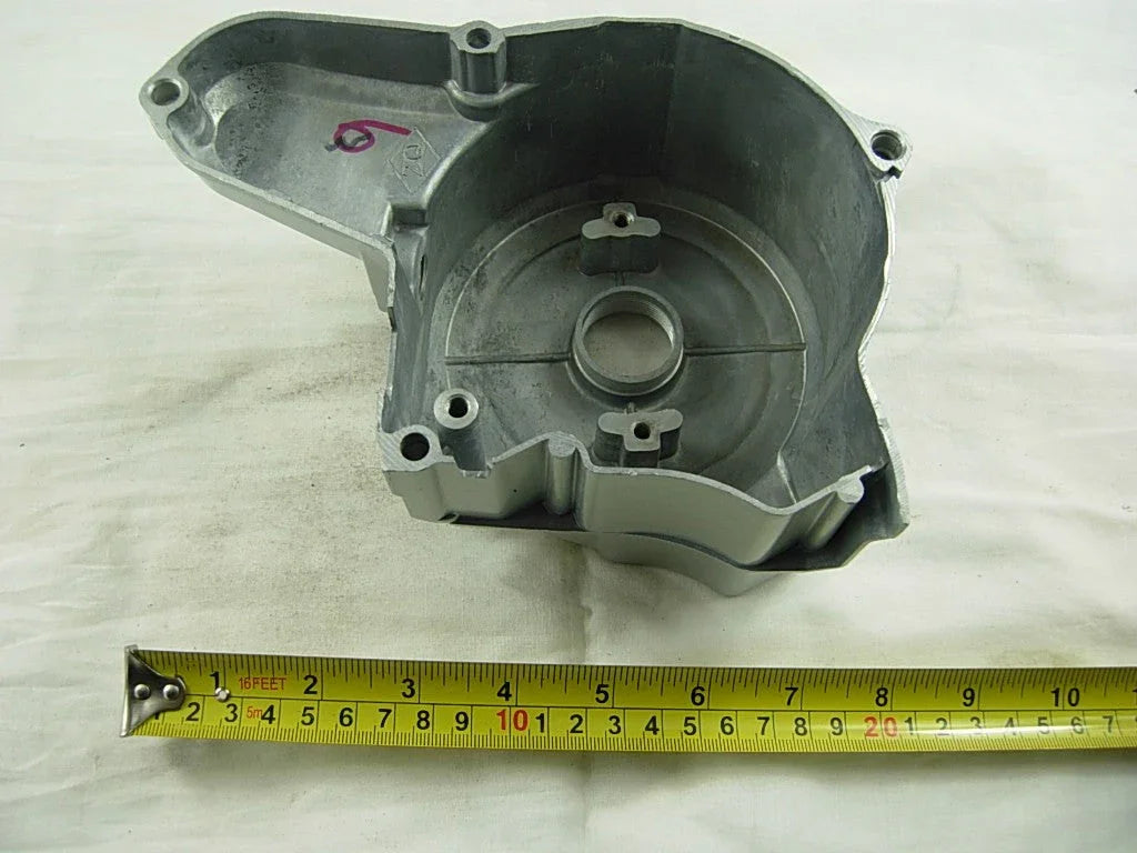 Metal engine timing cover part with mounting holes, placed on white surface next to a measuring tape