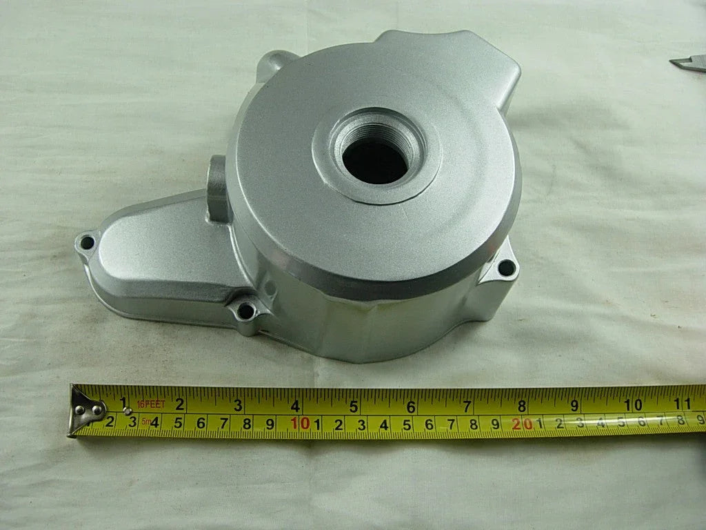 Silver metallic engine cover with circular opening placed on white surface next to yellow measuring tape