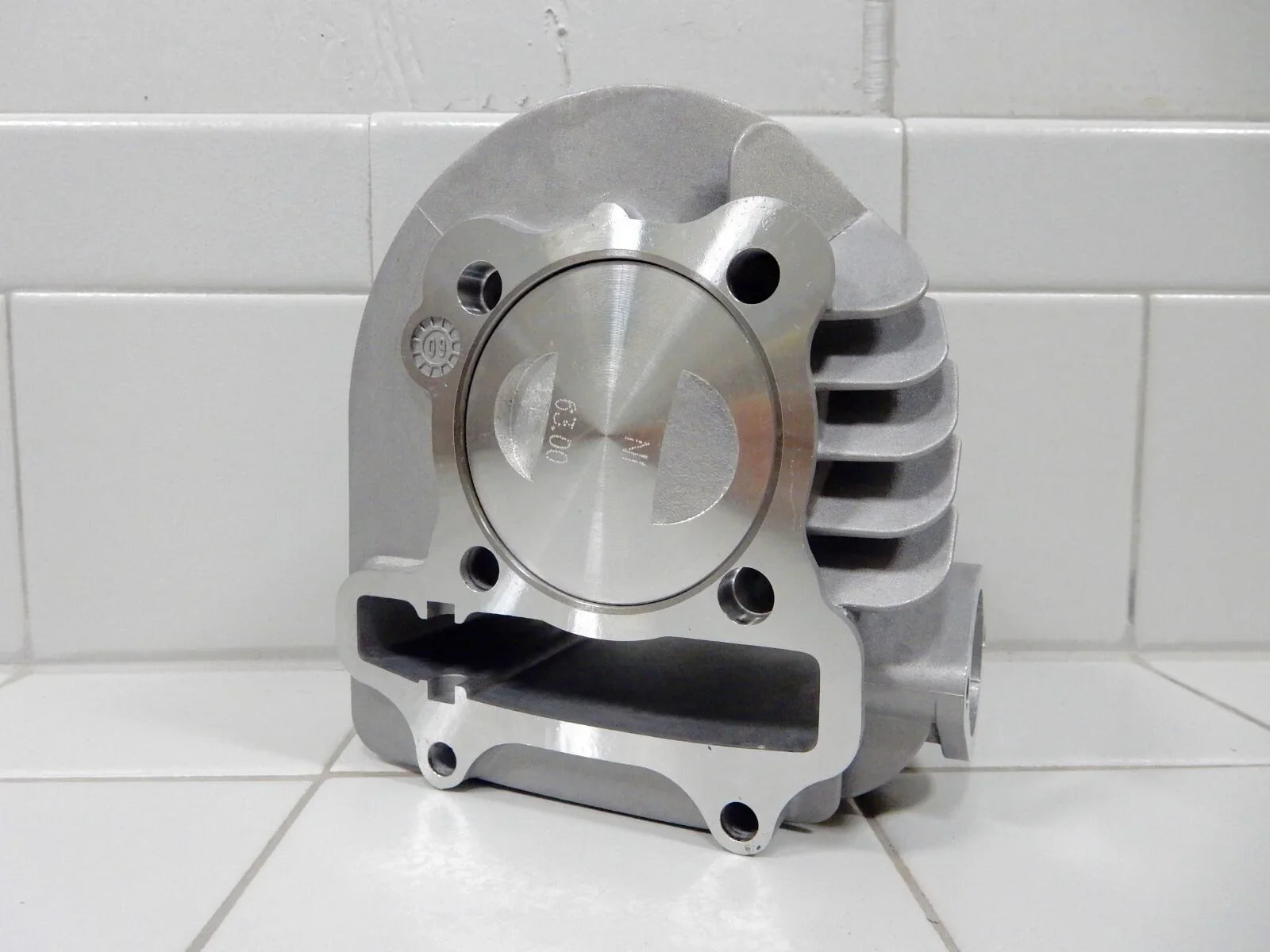 Silver motorcycle engine cylinder with piston marked 63.00 on white tiled surface