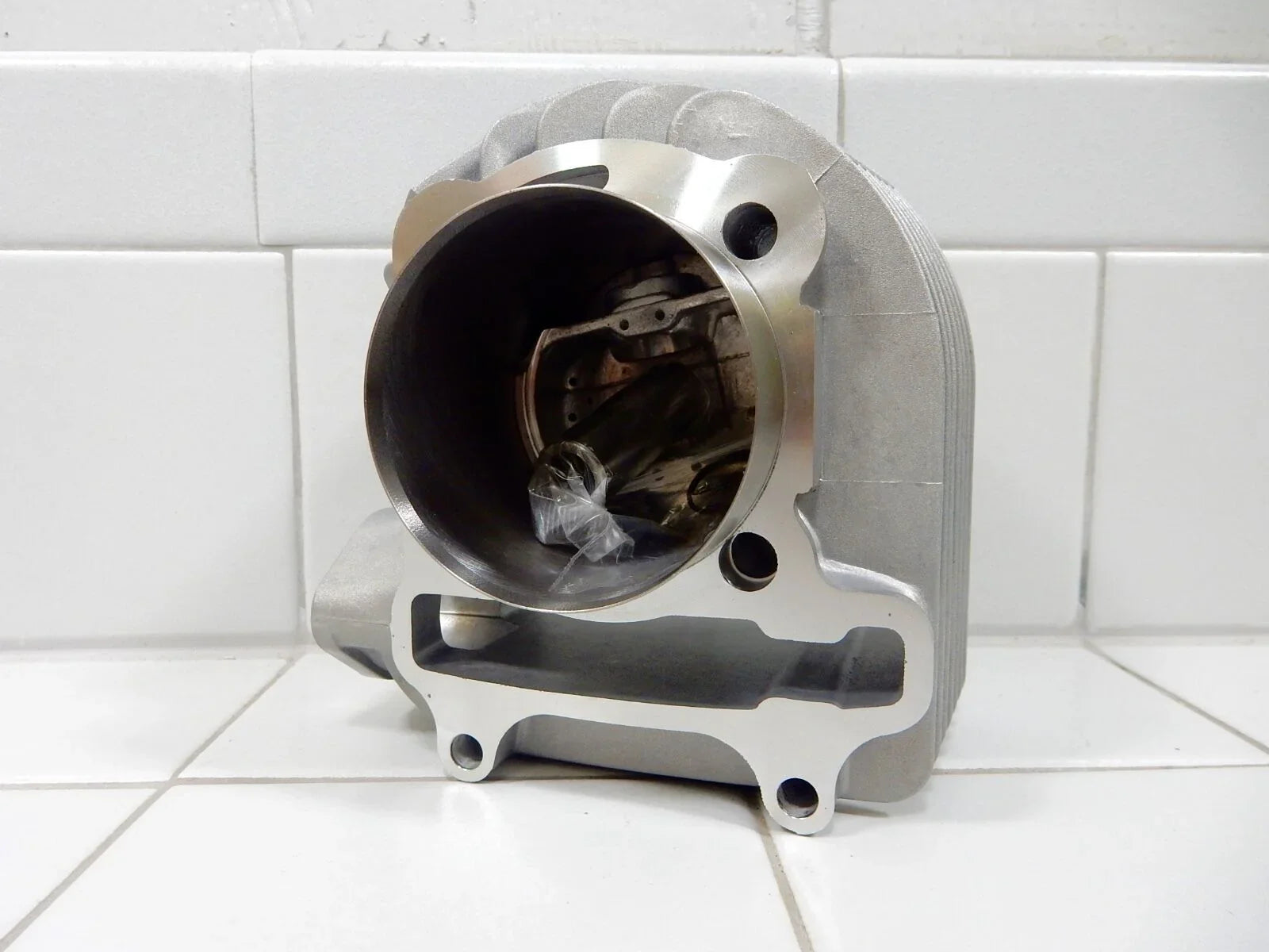 Aluminum motorcycle engine cylinder with visible piston pin inside, set on white tiled surface